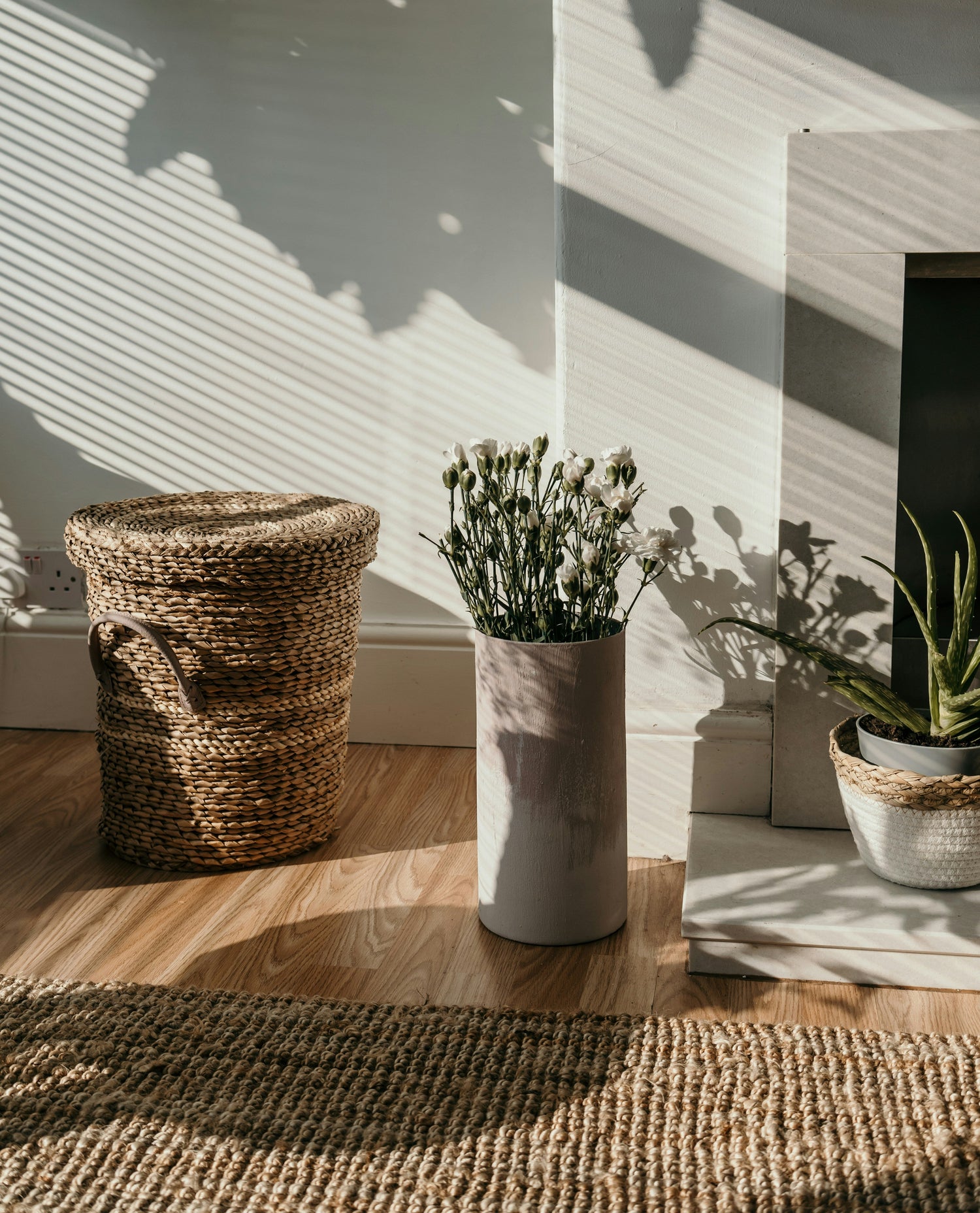 A woven basket, a ceramic floor vase with flowers, and indoor plants placed near a window in natural daylight.